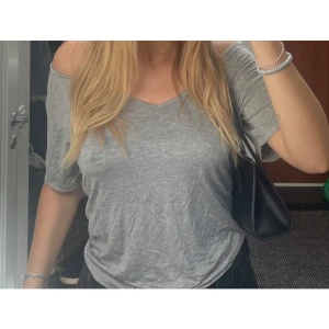 Grå offshoulder topp med axelband - Snygg grå offshoulder topp med kort ärm och unika tunna axelband i ryggen som korsar varandra. Toppens material är mjukt och lätt, perfekt för en chill och avslappnad stil. Djup v-ringning framtill och lös passform gör den extra bekväm.