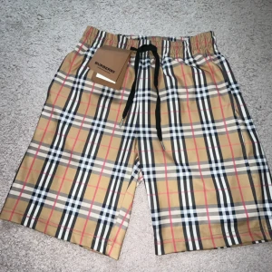 Burberry Shorts - Snygga Burberry shorts i klassiskt beige rutigt mönster med svarta, vita och röda detaljer. De har elastisk midja och svart snörning för perfekt passform. Perfekta för sommaren och ger en lyxig vibe till din outfit.