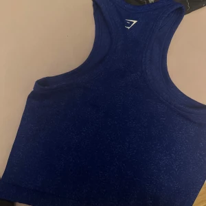 Blå linnetopp från Gymshark - Säljer en blå linnetopp från Gymshark med sportig racerback-design. Toppen är tillverkad i ett mjukt och stretchigt syntetmaterial som passar perfekt för träning. Snyggt melerat mönster och croppad passform som ger en modern look.