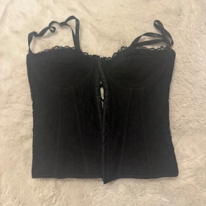 Svart spetskorsett med snörning - Säljer en svart korsett med spetsdetaljer och justerbara axelband. Framsidan har en snygg knäppning och baksidan har snörning.