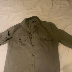 Olivgrön overshirt - Snygg olivgrön skjorta med klassisk krage och två bröstfickor med knappar. Skjortan har långa ärmar som kan rullas upp och fästas med knapp. Perfekt för en avslappnad men ändå stilren look.