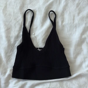Svart ribbad linnetopp från Zara - Svart ribbad linnetopp från Zara med smala axelband och djup v-ringning. Croppad modell som sitter tight och har en stilren look. Perfekt att styla med höga jeans eller kjol för en trendig vibe.