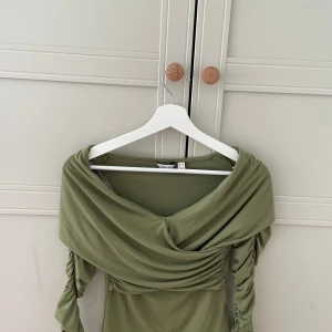 Olivgrön omlottopp från Vera Moda  - Snygg olivgrön omlottopp från Vera Moda  i storlek XS. Toppen har långa, rynkade ärmar och en draperad omlottdesign över axlarna. Mjuk och stretchig känsla, perfekt för en lyxig middag!