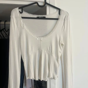 topp med spets från Gina Tricot - Supersöt vit  topp från Gina Tricot med långa ärmar, spetsdetaljer längs urringningen och en liten rosett framtill. Toppen har en lätt volang nertill. Använd 1 gång.