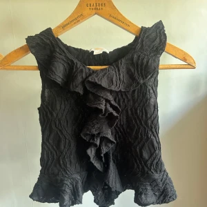 Svart volangblus från Gina Tricot - Supersnygg svart blus från Gina Tricot med volanger längs halsringningen och framtill. Toppens tyg är lätt transparent med struktur och har en vågig, cool känsla. Perfekt för dig som vill sticka ut lite extra med en trendig och unik topp.