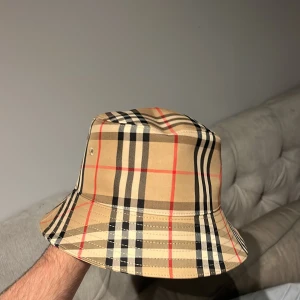 Burberry bucket hat i klassiskt rutmönster - Snygg bucket hat från Burberry i deras ikoniska beigea rutmönster med svarta, vita och röda detaljer. Hatten är tillverkad i bomull och har en mjuk, nedvikt brätte. Perfekt accessoar för att lyfta din stil.