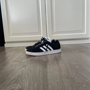”Grisch/skate” skor - Svarta Adidas Gazelle sneakers i mocka med klassiska vita ränder på sidorna och vit plös. Skorna har svart snörning och vit sula för en clean och tidlös look. Perfekta för dig som gillar streetstyle och vill ha något som funkar till det mesta.