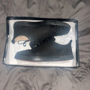 Svarta Louboutin sneakers med nitar - Säljer ett par svarta sneakers från Louboutin i mocka med ikoniska röda sulor. Skorna har svarta nitar på tån och svarta snören för en edgy vibe. Perfekta för dig som vill sticka ut med lyxig streetstyle. Skorna är använda några gånger men är i väldigt bra skick.