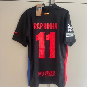FC Barcelona Raphinha #11 Nike tröja - Snygg svart FC Barcelona matchtröja med Raphinha och nummer 11 på ryggen. Tröjan har röda och blå detaljer, officiella loggor från Nike, Spotify och UEFA Foundation på ärmen. Tillverkad i lätt och ventilerande material, perfekt för fotboll eller att bära som supporter.
