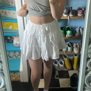 Vit volangkjol med snörning - Supersöt vit kortkjol med volanger och snörning i midjan. Kjolen har inbyggda shorts. Perfekt för varma dagar och lätt att matcha med crop tops eller linnen.