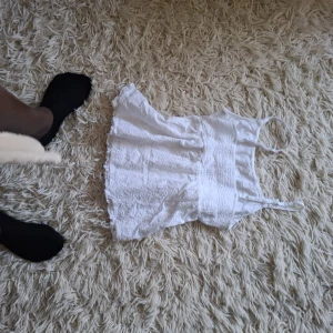 Vit broderad linnetopp med volang - Supersöt vit linnetopp med tunna axelband och broderade blommor över hela tyget. Toppen har smockad midja och volangkant nertill för en extra gullig look. Perfekt för varma dagar och ger en fräsch sommarkänsla.