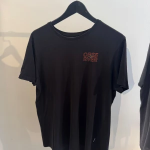 Svart t-shirt från Youturn - Cool svart t-shirt med trycket 'OBSERVER' i rött framtill och ett stort grafiskt tryck med öga och text på ryggen i rött och turkost. Klassisk rund hals och korta ärmar. Perfekt för dig som gillar streetstyle och unika prints.