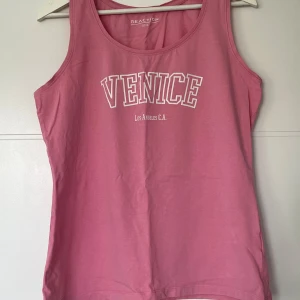 Rosa linnetopp med tryck från Reaction - Snygg rosa linnetopp från Reaction med vit text 'VENICE Los Angeles C.A.' framtill. Klassisk ärmlös modell med rund halsringning, perfekt för varma dagar. Mjuk bomullskänsla och avslappnad passform.