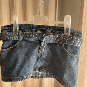 Jeanskjol med nitar från Pull&Bear XS - Snygg blå jeanskjol från Pull&Bear i storlek XS. Kjolen är kort och har ett brett bälte med silvriga nitar som ger en cool vibe. Klassisk femficksmodell och slitstarkt jeanstyg. Perfekt för dig som gillar edgy detaljer och denim.