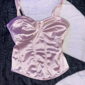 Ljusrosa satin korsett med rosett - Supersnygg ljusrosa korsettopp i glansig satin med tunna axelband och liten rosett framtill. Kuporna är formade för extra stöd och toppen har en figurnära passform som framhäver siluetten. Perfekt för dig som vill sticka ut med en trendig och feminin look.