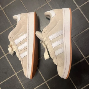Beige Adidas Campus sneakers mocka - Snygga beige Adidas Campus sneakers i mocka med klassiska vita ränder och vit häl. Skorna har en brun gummisula och vita snören. Perfekta för dig som gillar stilrena och tidlösa sneakers med retrovibbar.