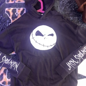 Nightmare Before Christmas hoodie - Svart hoodie från Disney med stor vit Jack Skellington-ansikte på bröstet och texten 'Jack Skellington' på ärmarna. Tryck med 'The Nightmare Before Christmas' på ryggen. Klassisk huva och magficka, perfekt för dig som älskar Tim Burton.