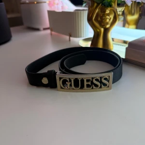 Svart skärp med guldspänne från Guess - Snyggt svart bälte från Guess med ett stort, rektangulärt guldspänne där märkesnamnet står ut. Bältet är smalt och stilrent, perfekt för att lyfta vilken outfit som helst. Tillverkat i skinn med elegant struktur och exklusiv känsla.