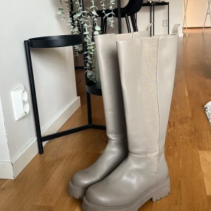 Beige höga boots med platåsula - Snygga höga boots i beige med chunky platåsula och rund tå. Skorna har elastiska paneler längs skaftet för en tight passform och är tillverkade i slätt syntetmaterial. Perfekta för att lyfta din outfit med en trendig touch.