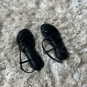 Svarta sandaler från VOX med remmar - Stilrena svarta sandaler från VOX med tunna remmar och justerbart spänne runt ankeln. Platt sula och minimalistisk design som ger en clean look. I strl 36 och använda max 2 gånger.