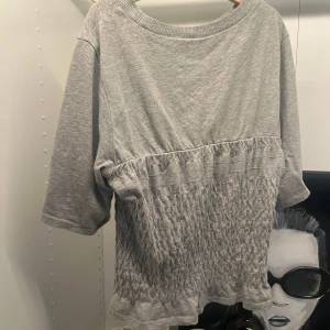 Grå topp med smockad midja - Grå topp i mjukt bomullsmaterial med trekvartsärm och bred rund halsringning. Nedre delen av tröjan har en smockad, rynkad effekt som ger en snygg och avslappnad look. Perfekt för dig som gillar en chill och trendig stil. Två sömmar har blivit lite lösare därav priset