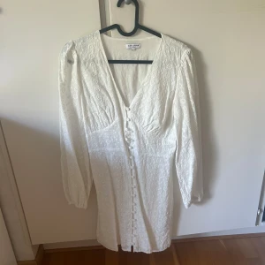 Vit spetsklänning från Bubbleroom - Superfin vit kortklänning från Bubbleroom med broderade blommor, v-ringning och knappar framtill. Långa ärmar med lätt puff och smock i ryggen ger en snygg passform. Perfekt för sommarens alla tillfällen!