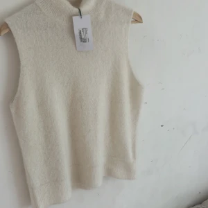 Vit stickad väst från Stockh LM Studio - Supermjuk och stilren vit stickad väst från Stockh LM Studio. Modellen är ärmlös med hög krage och ribbade kanter. Perfekt att bära över en skjorta eller t-shirt för en clean look. Materialet känns fluffigt och lyxigt. Original pris 599 med prislapp säljer för den är för stor