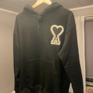 Ami Paris hoodie - Svart hoodie från AMI med stor vit och svart hjärta-A patch på bröstet. Klassisk huva med snörning och känguruficka framtill. Mjuk bomullskänsla och broderad AMI-logga i nacken. Perfekt för dig som gillar stilrena streetwear-plagg.