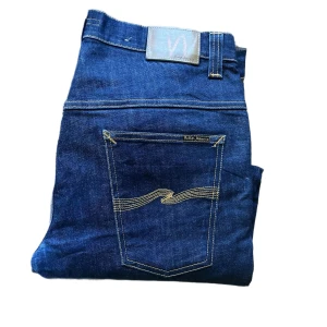 Nudie Jeans Tape Ted blå jeans - Säljer ett par mörkblå Tape Ted jeans från Nudie Jeans i klassisk femficksmodell. Jeansen har raka ben, snygga kontrastsömmar och är tillverkade i 99% ekologisk bomull och 1% elastan för lite stretch. Perfekta för dig som gillar stilrena och tidlösa jeans. Midja 44cm. Korta i storleken, kanske uppsydda tom men isåfall var dem det när jag köpte dom och isåfall väldigt bra uppsytt. Jag skickar ej bild när jag bär plaggen och jag vet ej vilken längd och vikt de passar då jag inte prövar kläderna 