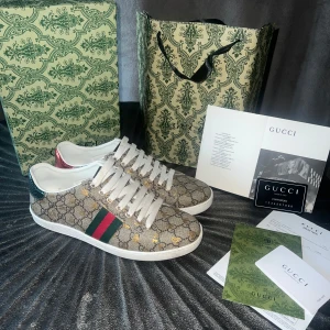 Gucci Ace sneakers med GG-mönster - Gucci Ace sneakers med klassiskt GG-monogram i beige canvas, gröna och röda ränder på sidan och metallic-detaljer i rött och grönt på hälen. Skorna har vita snören, guldfärgade bi-detaljer och vit platt sula. Perfekta för dig som vill sticka ut med lyxig streetstyle.