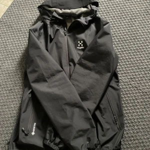 Svart Haglöfs Gore-Tex vindjacka - Svart vindjacka från Haglöfs i Gore-Tex-material. Jackan har huva, hel dragkedja framtill och två sidofickor med dragkedja. Perfekt för blåsiga och regniga dagar, med stilren design och diskret logga på bröstet.