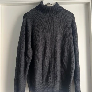 Mörkgrå polotröja från Belika L - Snygg mörkgrå polotröja från Belika i storlek L. Tröjan är stickad med ett diskret mönster och har en hög, ribbad polokrage. Materialet är en skön mix av 80% bomull och 20% ull, perfekt för kyliga dagar. Långärmad och med klassisk passform.