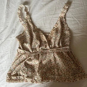 Blommig linnetopp med volangdetalj - Supersöt linnetopp i ljusbeige med små blommor i rosa och grönt. Toppen har omlott-skärning, smala axelband och volangdetaljer vid midjan och nederkanten. Perfekt för varma sommardagar och ger en romantisk vibe.