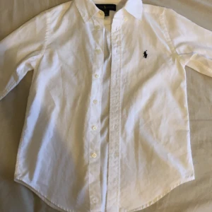 Vit skjorta från Ralph Lauren, strl 12 - Klassisk vit skjorta från Ralph Lauren med svart broderad logga på bröstet. Skjortan har lång ärm, knappar framtill och en snygg krage. Modellen är custom fit och tillverkad i bomull. Perfekt för en clean och stilren look.