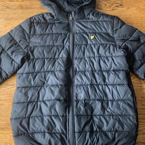 Svart dunjacka från Lyle & Scott - Svart dunjacka från Lyle & Scott med huva och dragkedja framtill. Jackan har horisontella sömmar, klassisk gul logga på bröstet och är perfekt för kyliga dagar. Enkel och stilren design som funkar till det mesta.