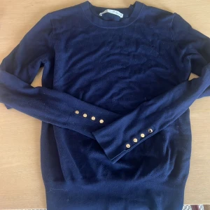 Mörkblå stickad tröja med guldknappar - Supersnygg mörkblå stickad tröja från Zara med rund hals och långa ärmar. Ärmsluten är dekorerade med fem guldiga knappar som ger en lyxig touch. Perfekt till jeans eller kjol för en stilren look. #stockholmstil