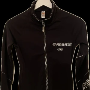 Svart gymnastikjacka från Agiva - Svart träningsjacka från Agiva med texten 'GYMNAST' tryckt på bröstet och ärmen. Jackan har lång ärm, hel dragkedja framtill med paljetter/diamanter på och smal passform. Perfekt för gymnastikträning eller uppvärmning. Storlek 10=128-140