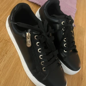 Svarta sneakers med gulddetaljer - Snygga svarta sneakers med vita sulor, guldfärgade öljetter och dragkedja på sidan. Skorna har snörning och är gjorda i ett slätt material som liknar skinn. Perfekta för en trendig och avslappnad look.