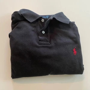 Svart pikétröja från Ralph Lauren - Klassisk svart pikétröja från Ralph Lauren med röd broderad logga på bröstet. Tröjan har korta ärmar, krage och två knappar framtill. Tillverkad i bomull för en skön och luftig känsla. Perfekt för en clean och stilren look.