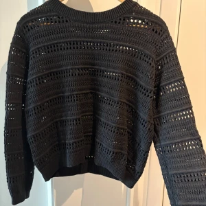 Svart stickad tröja med hålmönster - Snygga svart stickad tröja med lång ärm och coolt hålmönster i ränder. Tröjan har en relaxed fit och ribbade muddar vid hals, ärmslut och nederkant. Perfekt att styla över ett linne eller t-shirt för en trendig lager-på-lager-look.