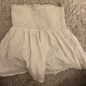 Vit kortkjol/topp från Gina Tricot - Supersöt vit kortkjol/topp från Gina Tricot med smockad midja och volangig nederdel. Kjolen är luftig och har en lätt rynkad struktur som ger en chill och somrig vibe. Perfekt att matcha med en crop top eller hoodie.