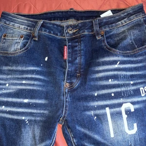 Dsquared2 blå jeans med tryck - Snygga blå jeans från Dsquared2 med coolt vitt tryck och slitningar. Jeansen har en tvättad look med färgstänk och klassisk femficksmodell. Raka ben och normal passform, perfekt för dig som gillar streetstyle och detaljer som sticker ut. 