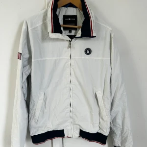 Vit vindjacka från Sail Racing L - Vit vindjacka från Sail Racing med dragkedja, ståkrage med marinblå och röda detaljer samt amerikansk flagga på ärmen. Jackan har två fickor framtill och broderad logga på bröstet och på ärmarna. 1 innerficka. Perfekt för dig som gillar sportig och stilren look.