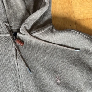 Ralph Lauren zip hoodie - Snygg grå hoodie från Polo Ralph Lauren med dragkedja, huva och snörning. Lite annorlunda från deras vanliga zip hoodies, denna är i högre kvalitet, det syns på den första bilden. Den är ganska liten i passformen så skulle säga att den sitter mer som en Small även fast det är en Medium. Tveka inte att höra av dig vid minsta lilla fråga!