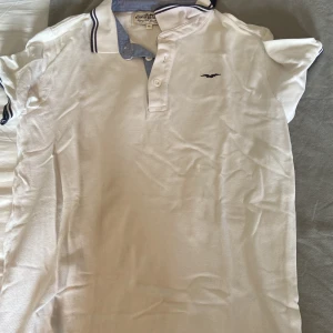 Vit pikétröja Hampton Republic 170 - Vit pikétröja från Hampton Republic med klassisk krage och blå detaljer på insidan av kragen. Tröjan har korta ärmar, knappar framtill och en liten broderad logga på bröstet. Perfekt för en clean och stilren look. Storlek: 170, skick: 8/10, kontakta vid frågor!