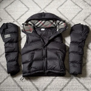 Burberry jacka/väst  - Snygg svart jacka/dunväst från Burberry med avtagbara ärmar. Västen har dragkedja, tryckknappar och två fickor framtill. Insidan av huvan har klassiskt rutigt Burberry-mönster i grått o svart. Perfekt för lager-på-lager och streetstyle.