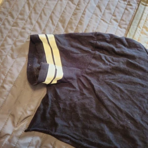 Svart croppad t-shirt Divided S - Svart croppad t-shirt från Divided med korta ärmar och två vita ränder på ärmsluten. Klassisk rund halsringning och loose fit. Perfekt för en chill och sportig vibe.