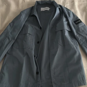 Blå overshirt från Calvin Klein - Snygg blå overshirt från Calvin Klein med klassisk krage, två stora bröstfickor och svarta knappar framtill. Jackan har en stilren look och patch med logga på ärmen. Perfekt för lager-på-lager och enkel att matcha. Aldrig använd