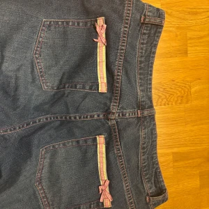 Vintage Lilly Pulitzer jeans med rosetter - Mörkblå jeans från Lilly Pulitzer med raka ben och låg midja. Snygga detaljer med rosa och gula ränder samt små rosetter på bakfickorna. Jeansen har rosa sömmar och är tillverkade i bomull med lite stretch för extra komfort. Pris kan diskuteras!!