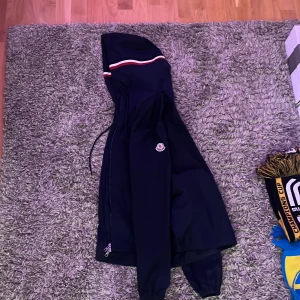 Mörkblå vindjacka från Moncler - Säljer en mörkblå vindjacka från Moncler med huva och snygga röda och vita ränder på huvan. Jackan har en liten skada som man ser på bild 4 och en liten fläk vid dragkedjan 